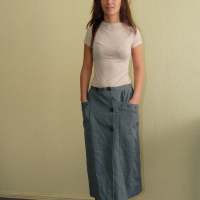 A person stands indoors against a light green wall. They have long brown hair and are wearing a fitted beige t-shirt and a high-waisted, ankle-length green skirt with a front button detail and pockets. Their black sandals contrast with the dark floor material, reflecting modern Modes of fashion.