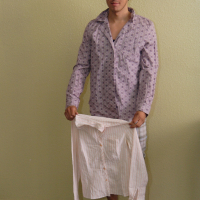 A young man stands indoors against a light green wall wearing sunglasses, an open-collared long-sleeve patterned shirt, and striped shorts. He is holding another long-sleeve shirt, which is white with thin vertical stripes, in front of him with both hands. His black and white striped slip-on shoes add to his modes talka flair.