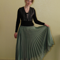 A person stands against a yellow wall, wearing a black long-sleeve shirt with a sleeveless, black leather-look vest from Talka Modes, and a flowing, pleated green skirt. They hold the skirt delicately with one hand and wear black lace-up boots. Their hair is pulled back, and they are smiling slightly.