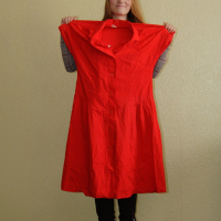 A person with long hair is standing against a light-colored wall, holding up a bright red dress by the shoulders, showcasing it as if in a Modes Talka photoshoot. The dress features a button-down design. Wearing a grey checkered shirt and dark pants, black boots peek out at the bottom.