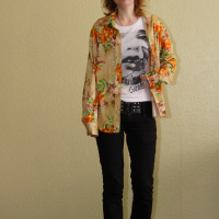 A person with shoulder-length hair is standing indoors against a light-colored wall. They are wearing a bright floral shirt over a graphic t-shirt, black pants, and black shoes. Adopting a relaxed mode, they look straight ahead with a neutral expression and have one hand partially in a pocket.