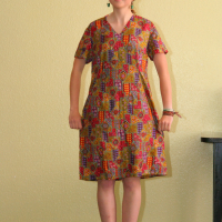 A woman stands indoors against a beige wall, wearing glasses and a colorful, patterned dress that reaches her knees. She is also wearing red sneakers and has her hands relaxed by her sides. Her hair is tied back, and she has a slight smile on her face. A partial view of a Modes Talka painting is visible above her.