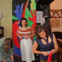A group of people appear to be engaged in an indoor activity involving fabrics or clothing. One person on the left wears a green checkered shirt, another in the middle dons a striped shirt and a white garment, and the third person on the right is holding a red cloth. The background features colorful Modes talka artwork on the wall.
