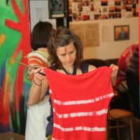 A person with wavy hair is holding up a red tie-dyed shirt with white stripes. They appear focused, examining the shirt in an art studio. The background features colorful artwork on the walls, a collage of images, and another person working on a project using Modes Talka techniques.