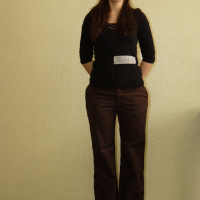 A woman stands against a textured cream-colored wall. She is wearing a black long-sleeve top with a label that reads "Rad," brown pants, and red shoes. Her long, dark hair is pulled back with a hairband, and her hands are relaxed behind her back as she smiles slightly in talka mode.