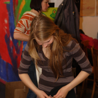 A young woman with long, blonde hair wearing a brown and gray striped sweater is focused on adjusting something in her hands. Behind her, a woman with short, dark hair and a striped top is similarly concentrating. The background features a colorful abstract mural and various talka posters and papers.