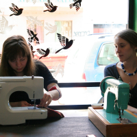 Two women are sitting at a table with sewing machines in a cozy room. The woman on the left is focused on sewing fabric with a modern white machine, while the woman on the right observes, her vintage teal machine in front of her. Modes talka bird decals adorn the large window behind them, allowing sunlight to stream in.