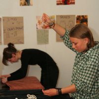 Two people are engaged in sewing activities in a room decorated with posters and notes on the wall. The person in the foreground, wearing a green checkered shirt, is holding a thread, while the person in the background leans over fabric, working with various sewing tools scattered on the table. Their focused modes of work create a harmonious scene.
