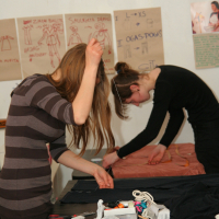 Two women are working on fabric at a table cluttered with sewing supplies like scissors, threads, and pins. They are both focused on their tasks, with one threading a needle and the other bending over her work. In the background, sketches and notes on Modes talka fashion designs are pinned to the wall.