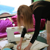 A person with long hair is bending over a table, working with a polka dot fabric. The room, brightly lit by a large window revealing part of a blue car outside, has a sewing machine on the table to their left and some pink Modes Talka fabric in the background.