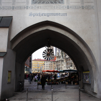 An arched entrance with the sign "spielzeugmuseum" leads to a vibrant city square in Munich, where a colorful market is visible and a historic building with intricate façades stands in the background