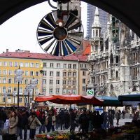 An archway frames a bustling Munich city square where vendors sell vibrant flowers beneath a historical clock, with gothic architecture and urban facades blending in the backdrop.