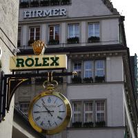 A golden Rolex sign with an ornate clock extends over the sidewalk in front of a multi-story building with the name "Hirmer" on its facade, suggesting a luxury watch retailer in an urban