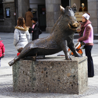 A person interacts with a bronze boar statue in a bustling Munich city square, where passersby, including a child in a red jacket, go about their day.