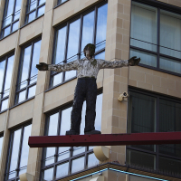 A life-size sculpture of a human figure with arms outstretched stands on a red beam, mounted on the facade of a modern building in Munich with large windows and stone cladding, creating an eye