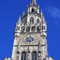 A clear blue sky provides a backdrop for the ornate, gothic tower of a historic building in Munich, featuring intricate spires, a large clock face, and statues nestled in its architecture.