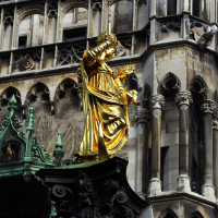 A gilded statue gleaming in the sunlight, possibly a historical or religious figure, stands before an ornate gothic building with detailed stone facades and architectural decorations in Munich.