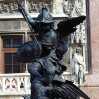 A bronze statue of a warrior angel, wielding a sword and shield, stands defiantly with wings outstretched against a backdrop of ornate gothic architecture in Munich.