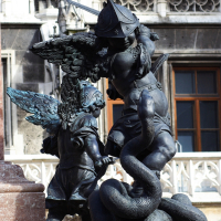 A bronze statue depicting a winged warrior engaged in a fierce battle against a serpentine monster, all set against the backdrop of an ornate building with classical architectural features in Munich.