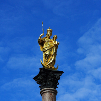 A golden statue of a robed female figure holding a spear and shield, standing atop a tall column against a clear blue sky in Munich.