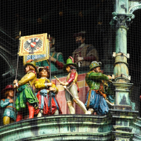 An array of colorful, historic figures displayed behind a protective mesh in Munich, possibly a detail from an ornate, mechanical clock tower, where intricately dressed sculptures prepare to enact a traditional hourly show.