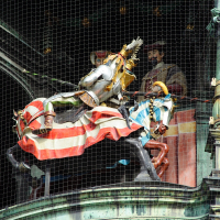 A vibrant, medieval knight statue with a golden lance on horseback, accompanied by figures in period attire, all ensconced behind protective netting atop an ornate, green-roofed structure in