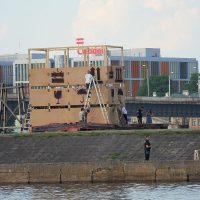 Several people are assembling a large wooden structure resembling part of a building facade along a riverside during the Kapusvētki event. Two individuals are on ladders adjusting the structure. In the background, modern buildings are visible. One person stands near the waterway, with greenery in the distance.