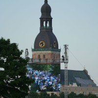 A tall, dome-topped church tower with a weather vane rises against a clear blue sky. Below, a large screen shows a crowd of people, possibly at Kapusvētki. The setup is outdoors, with tree foliage and scattered onlookers in the foreground. The scene conveys a blend of historic architecture and modern technology.
