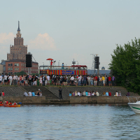 A crowd gathers at a riverside pier for the Kapusvētki event featuring a stage with colorful decorations. A red kayak and a white motorboat with people in orange vests are visible in the water. In the background, an imposing historic building with a tall spire stands against a light blue sky.