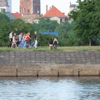 A group of people walks along a riverside path bordered by trees and historic buildings, reminiscent of a Kapusvētki gathering. One individual rides a bicycle while another sits on a bench, gazing at the serene river water. It's a bright, sunny day with clear skies.