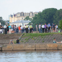 A group of people, including police officers, are gathered on a promenade by a river with historical buildings and trees in the background. During the Kapusvētki event, some individuals sit on the stone steps leading to the river, engaging in conversation, while others stand near bicycles or in groups.