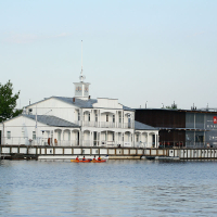 A serene waterfront scene showcases a white, multi-story boathouse with a clock tower and veranda. Beside it, a dark, modern building with "BRIGANTIA" signage stands tall. Two people in a red-orange kayak paddle on the calm water in the foreground, enjoying the tranquility of Kapusvētki. Trees flank the buildings under a clear sky.