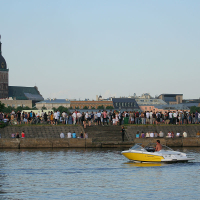 A yellow and white speedboat with a single person on board cruises along a river in the foreground. In the background, numerous people are gathered along the riverbank near a historic church with a tall clock tower, celebrating Kapusvētki. The sky is clear and light blue, indicating a calm and pleasant day.