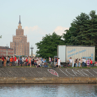 A crowd of people, including some with bicycles, gathers by the water's edge in an urban area during Kapusvētki. A truck with a blue banner is parked nearby. In the background, a tall, ornate building with a spire is visible. Graffiti on the embankment reads "I can't live" with a heart symbol next to it.