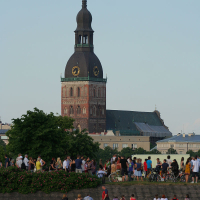 A crowd of people gathers near a riverbank, with some sitting on the grass and others on bicycles. In the background stands a historic church with a prominent dark spire and clock faces, reminiscent of Oligarhu kapusvētki celebrations. The sky is clear, and the scene is bustling with activity.