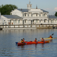 Four people paddle kayaks on a calm river in front of a white, two-story boathouse with "Waiska" written on the side. The scene, perhaps part of kapusvētki celebrations, is set in daylight with trees and other buildings in the background.