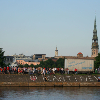 A large crowd gathers along the riverside embankment during Kapusvētki. In the background, historic buildings and a tall church spire rise into the blue sky. Graffiti on the embankment reads "YOU + ME = ♥ I CAN'T LIVE WITHOUT YOU." Trees frame the scene on both sides.