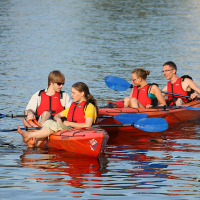 Four people in red life jackets sit in two tandem kayaks on calm water. Each kayak has one man and one woman. They are holding blue paddles and appear to be talking and laughing, enjoying their time on the water during kapusvētki. The scene is set in daylight with reflections visible on the surface.
