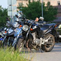 Several parked motorcycles with various designs and models, including a Honda, are lined up on the side of the road near a grassy area. In the background, an old vehicle is seen, and two people are near the motorcycles. The scene resembles a festive kapusvētki gathering with buildings and greenery visible in the distance.