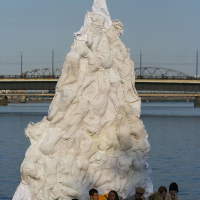 A group of people sit next to a river beneath a large, white, conical art installation made of fabric. Reminiscent of the Oligarhu kapusvētki celebration, the structure resembles a tree or mountain and is held up by a central pole. In the background, a bridge spans across the river under a clear blue sky.