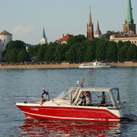 A small red and white boat with four people on board cruises along a calm river during Kapusvētki, with a European cityscape in the background. The city features historic buildings, green trees, and several tall church spires. Another boat is seen in the distance. The sky is clear, suggesting a pleasant day.