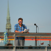 A man with a beard and sunglasses stands at a podium on an outdoor stage during the Oligarhu kapusvētki, speaking into a microphone while holding papers. He is wearing a blue plaid shirt. The stage is decorated with colorful leis, and a tall, pointed structure is visible in the background under the clear blue sky.