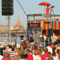 A colorful double-decker bus serves as an outdoor stage at a riverside event with a crowd seated on grass. One person is filming from the top of the bus, while another speaks into a microphone. Against a backdrop of city buildings and a bridge, someone holds an Oligarhu protest sign, evoking the spirit of kapusvētki.