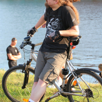 A person with long hair sits on a gray bicycle near a riverbank during kapusvētki. They wear sunglasses, a black T-shirt with a dragon design, gray shorts, gloves, and black shoes with white socks. Two other people stand nearby looking out over the water. The background includes more people and a green lawn by the water.