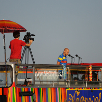 A man in a striped shirt speaks into a microphone under a blue sky atop a colorful double-decker bus adorned with flowers, celebrating the lively spirit of Kapusvētki. Beside him, a cameraman in a red shirt records from under a large rainbow-colored umbrella. The bus features vibrant horizontal stripes and the website "SHOWbus" is visible.