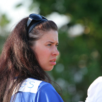 A woman with long, dark hair stands outdoors with a thoughtful expression during Kapusvētki. She is wearing a blue and white shirt, and sunglasses are perched on her head. The green, blurry background indicates a lush setting. Only the side and back head and shoulders of another person are visible.