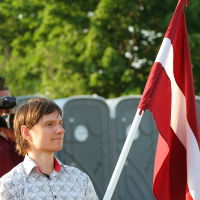 A person with short hair is standing outdoors, holding a large Latvian flag with a red field and white horizontal stripe. They are wearing a patterned short-sleeve shirt, possibly attending the kapusvētki festival. Behind them are portable toilets and trees with green leaves, under a clear sky indicating bright weather.