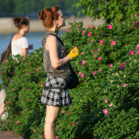 A woman with red hair, tied in a bun, wearing a leopard print sleeveless top, black and white plaid skirt, and yellow gloves, closely examines a bush with pink flowers. She stands on a paved path with her bag and other belongings at her feet. In the background of this serene scene resembling an oligarhu kapusvētki, a body of water and trees can be seen