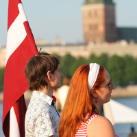 A young man and woman stand side by side near a red and white flag during kapusvētki. The man, in a patterned shirt, has short brown hair. The woman, with long red hair, wears a white headband and red and white striped top. A historical building with a large tower is blurred in the background, along with trees and water.