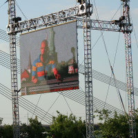 A large outdoor screen is mounted on a metal frame structure with wires for support. The screen shows a person in a multicolored striped shirt speaking at the Oligarhu Kapusvētki festival. The setup is in a grassy area with small trees, and a body of water can be seen in the background under a clear sky.